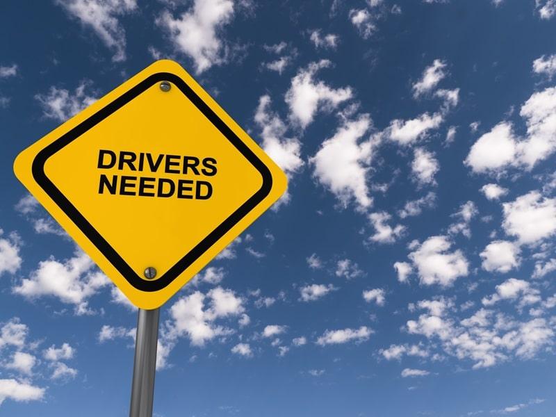 A yellow US-style road sign with "Drivers Needed" written on it